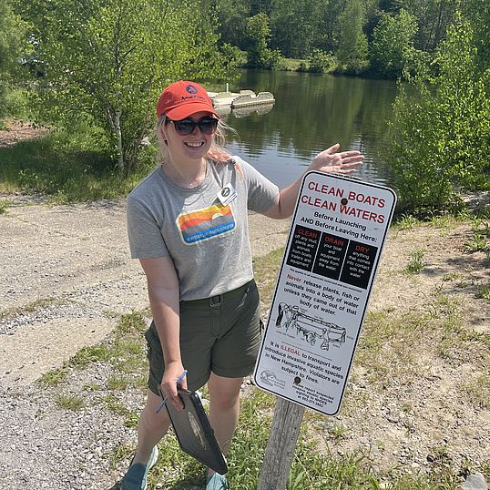 A smiling lake host indicates a “Clean Drain Dry” sign.
