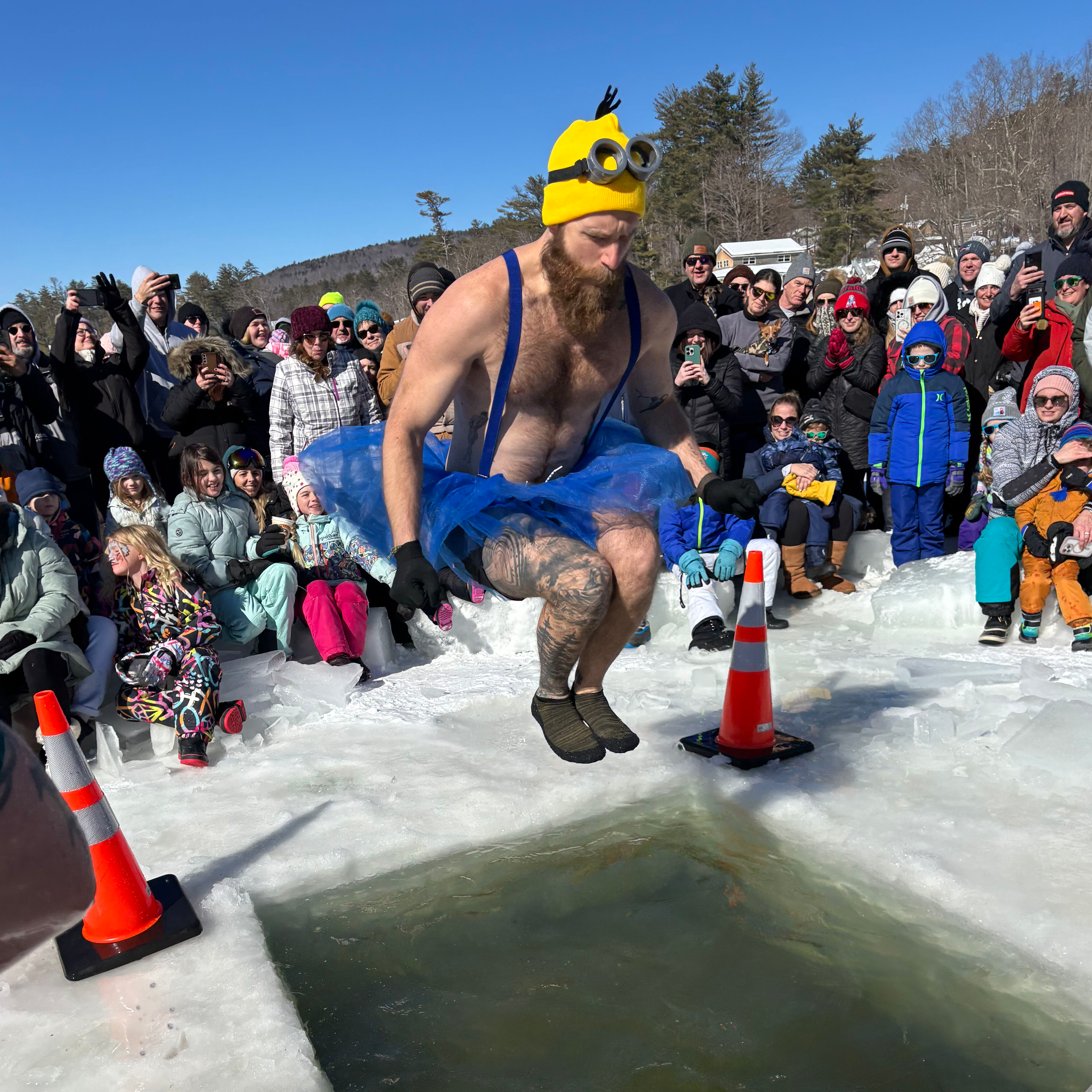 A costumed participant jumps through a hole in the ice with a large crowd looking on.
