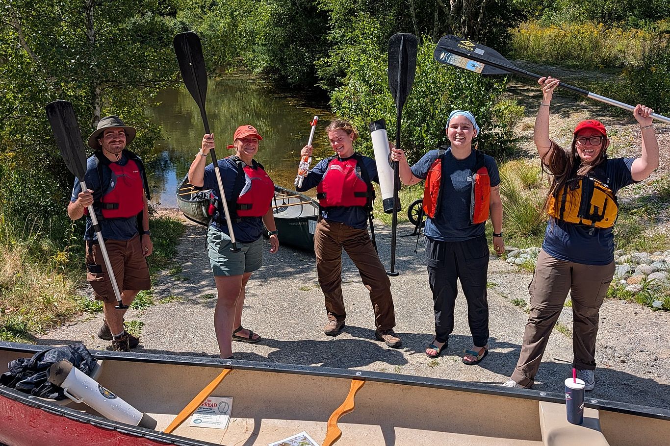Five weed watchers stand behind a canoe in preparation for the weed stampede, with paddle and equipment raised in a celebratory pose.